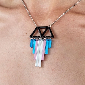 Könnte beinhalten: Halskette mit silberner Kette und schwarzem geometrischen Anhänger. Der Anhänger hat ein dreieckiges Design mit herabhängenden blauen, rosa und weißen rechteckigen Akzenten. Die Halskette wird auf der Brust einer Person getragen.