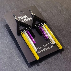 May include: Dangle earrings featuring black triangular shapes and vertical bars in yellow, silver, and purple. The earrings are presented on a black card with the text "Maine + Mara" and "maineandmara.com".