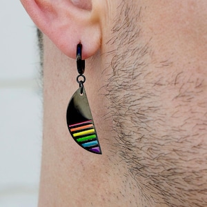 May include: A black earring with a half-moon shape and a rainbow stripe design. The earring is attached to a black hoop. The rainbow stripes include red, orange, yellow, green, blue, and purple.