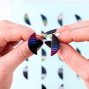 Peut inclure: Boucles d'oreilles noires semi-circulaires avec des rayures horizontales roses, violettes et bleues. Les boucles d'oreilles sont tenues entre deux mains sur un fond bleu clair avec des motifs similaires.