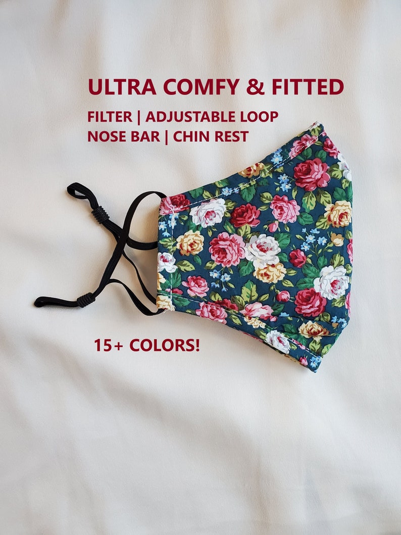May include: A floral face mask with a dark teal background and a pattern of pink, red, and yellow roses. The mask has adjustable black ear loops and a nose bar. Text on the mask reads: ULTRA COMFY & FITTED, FILTER | ADJUSTABLE LOOP, NOSE BAR | CHIN REST, and 15+ COLORS!