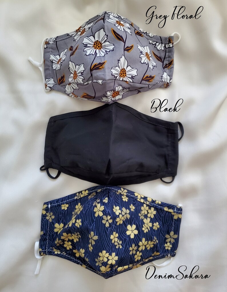 May include: Three face masks in different patterns. The top mask is grey with white floral print, the middle mask is solid black, and the bottom mask is navy blue with gold floral print. The text "Grey Floral", "Black", and "Denim Sakura" are written on the image.