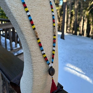 May include: A long beaded necklace with a colorful pattern of red, yellow, blue, black, and white beads. The necklace has wooden beads and a large dark brown pendant with a red tassel. The necklace is displayed on a mannequin.