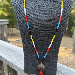 May include: A beaded necklace with a colorful pattern of red, blue, yellow, and white beads. The necklace has a large, round, brown pendant with a red tassel. The necklace is displayed on a black mannequin.
