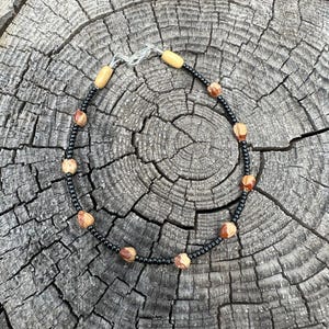 May include: Ankle bracelet featuring a black beaded chain with alternating light brown and tan beads. The bracelet also includes small, light brown cylindrical beads and a silver-colored clasp. The bracelet is displayed on a weathered, gray wooden surface.