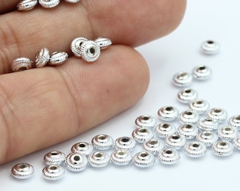 May include: A close-up of a hand holding a pile of silver metal beads. The beads are round with a raised edge and are scattered on a white surface.