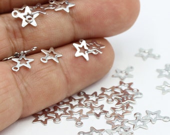 Puede incluir: Primer plano de una mano sosteniendo un montón de dijes en forma de estrella plateados con anillos de salto. Los dijes están esparcidos sobre una superficie blanca.