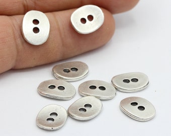 May include: A group of ten silver-toned, oval-shaped buttons with two holes each. The buttons are scattered on a white surface.