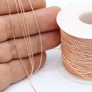 May include: A spool of rose gold colored chain, the chain is wound around a white spool. The chain is also shown laid out on a hand.