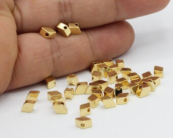 May include: A collection of small, gold-colored, geometric beads with a square shape and a hole through the center. The beads are scattered on a white surface, with some held in a hand.