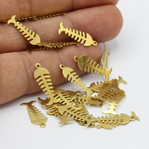 May include: A pile of gold-colored metal fish bone charms. The charms are about 1 inch long and have a loop at the top for attaching to jewelry or other crafts.