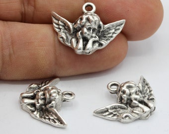 Puede incluir: Tres encantos de ángel de plata con alas y una pose de oración. Los encantos están sobre una superficie blanca.