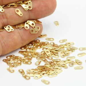 May include: Small, gold-coloured metal connectors with two holes and a rectangular shape are scattered on a white surface. These jewellery findings are used for connecting pieces in jewellery making and other crafts.