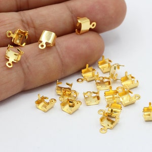 May include: A group of gold-toned metal clasps with a square shape and a loop on each side. The clasps are scattered on a white surface.