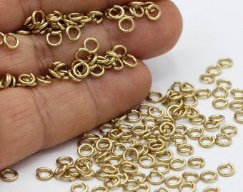Puede incluir: Una gran cantidad de pequeños anillos de salto de color dorado. Los anillos están esparcidos sobre una superficie blanca y se sostienen en una mano.