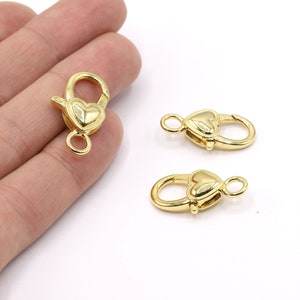Könnte beinhalten: Drei goldfarbene Herz-Lobster-Verschlüsse. Die Verschlüsse sind etwa 2,5 cm lang und haben einen federbelasteten Mechanismus.