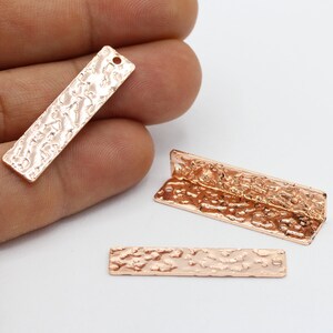 May include: Three rose gold metal rectangular charms with a hammered texture. Two charms have a hole at the top for hanging. The third charm has a corner cut out.