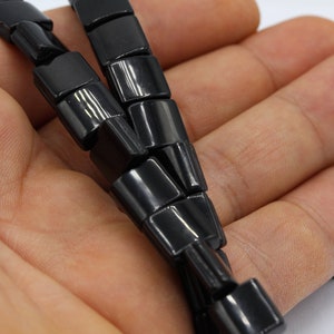 May include: A black beaded necklace made of square-shaped beads. The necklace is lying on a person's hand.