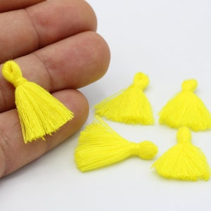 May include: Six bright yellow fabric tassels, each with a small loop at the top. The tassels are arranged in a semi-circle on a white surface.