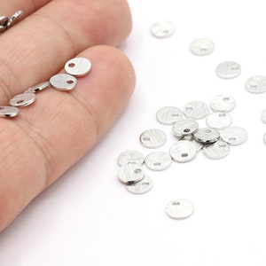 May include: Small, silver-colored, round metal charms with a textured surface and a small hole. These charms are scattered on a white surface, with some held in a hand. Ideal for jewelry making or crafting.