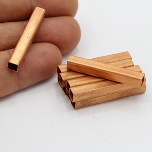 Pode incluir: Uma pilha de tubos quadrados de cobre, com uma peça sendo segurada na mão. Os tubos são de cor cobre quente e têm um acabamento brilhante.