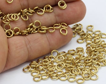 Puede incluir: Un montón grande de anillos de salto dorados, también conocidos como anillos partidos, están esparcidos sobre una superficie blanca. Los anillos son pequeños y redondos, y son perfectos para conectar piezas de joyería.