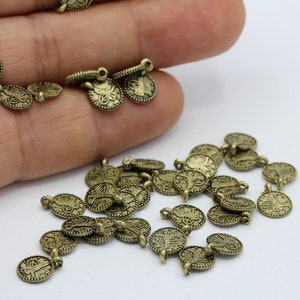Könnte beinhalten: Eine große Gruppe antiker goldfarbener Metallanhänger mit einem dekorativen Design. Die Anhänger haben oben eine Öse zum Anbringen an Schmuck oder andere Bastelarbeiten.