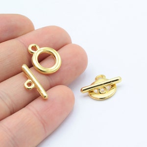 May include: Two gold-toned metal toggle clasps. One clasp is a round ring with a small loop at the top. The other clasp is a flat bar with a round ring in the center.
