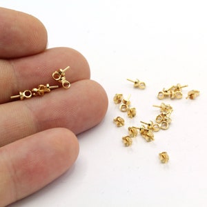 Puede incluir: Primer plano de un montón de hallazgos de pendientes de color dorado. Los hallazgos son pequeños y tienen un bucle en la parte superior para sujetar una cuenta u otro componente de joyería.