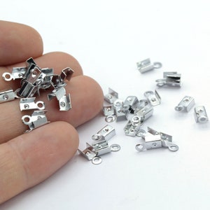 Puede incluir: Un montón de cierres de metal plateado con un bucle en un extremo y un borde plano en el otro. Los cierres están dispersos sobre una superficie blanca.