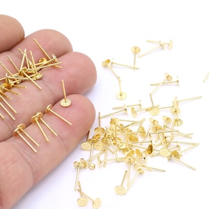 May include: A large pile of gold-coloured earring posts with flat round tops. The posts are scattered on a white surface.