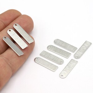 Op de afbeelding: Een set van acht zilverkleurige metalen labels met een geborstelde afwerking. De labels zijn rechthoekig met afgeronde hoeken en hebben een gat aan de bovenkant om een ​​touwtje of lint aan te bevestigen.