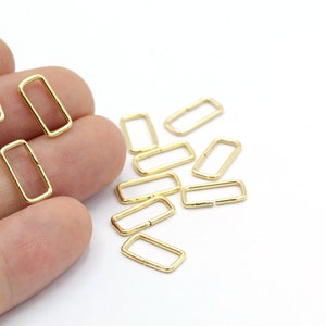 May include: A group of gold-colored metal rectangular rings, also known as D-rings, are scattered on a white surface. The rings are about 1 inch long and 1/2 inch wide.