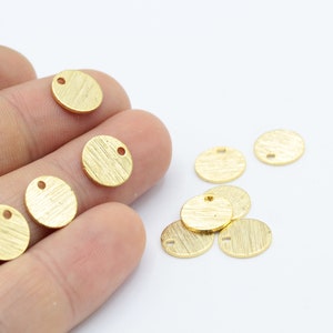 May include: A collection of small, round, gold-colored metal charms with a hole in the center. The charms have a textured surface and are scattered on a white background.