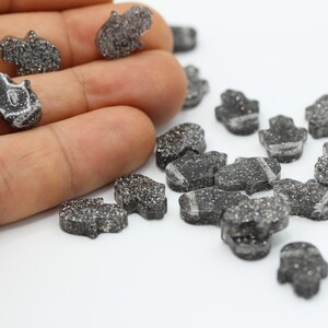 May include: A collection of small, black, glittery hamsa hand charms. The charms are scattered on a white surface.