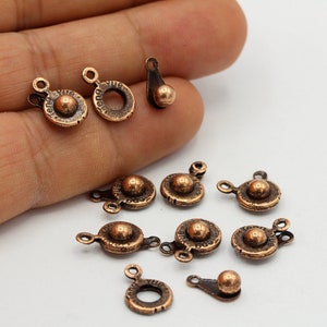 Puede incluir: Una colección de pequeños abalorios de metal de color bronce antiguo. Los abalorios presentan un diseño circular con una esfera central elevada y un pequeño lazo para la fijación. Algunos tienen un pequeño elemento de bola separado.