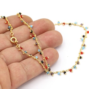 Op de afbeelding: Een gouden ketting enkelbandje met kleine, kleurrijke kralen die eraan hangen. De kralen zijn rood, blauw, zwart en helder.
