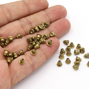 Pode incluir: Uma coleção de pequenas costas de brincos de cor latão, espalhadas sobre uma superfície branca. As costas dos brincos têm uma forma cónica com um pequeno orifício no centro. A imagem é bem iluminada, destacando o brilho metálico das costas dos brincos.