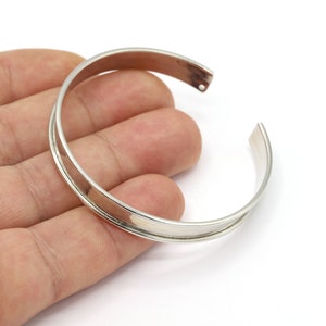 May include: A silver bangle bracelet with a wide, flat band. The bracelet is open and has a simple, minimalist design.