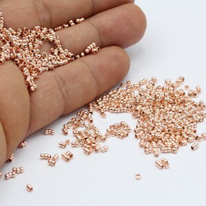 May include: A large pile of small, rose gold colored metal beads. The beads are round and have a smooth finish. They are scattered on a white surface.