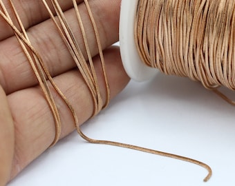 May include: A spool of gold-colored snake chain, a type of jewelry chain, is shown. The chain is wound around a white spool. A length of the chain is laid out on a hand.