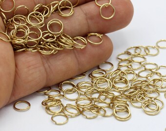 Puede incluir: Un montón grande de anillos de salto dorados, un tipo de hallazgo de joyería, están esparcidos sobre una superficie blanca. Algunos de los anillos se sostienen en una mano.