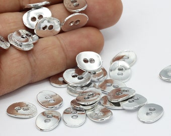 May include: A pile of silver oval-shaped buttons with two holes each. The buttons are scattered on a white surface.