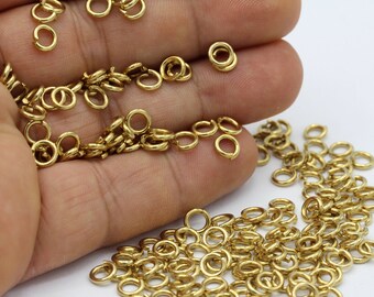 Puede incluir: Un montón grande de anillos de salto dorados, también conocidos como anillos partidos, están esparcidos sobre una superficie blanca. Los anillos son pequeños y redondos, y son perfectos para conectar piezas de joyería.