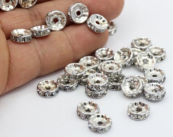 May include: A close-up of a pile of silver-toned metal bead caps with clear rhinestones. The bead caps are round and have a flat back.