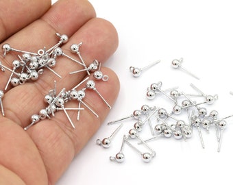 May include: A pile of silver earring posts with small round balls on the ends. The earring posts are scattered on a white surface.