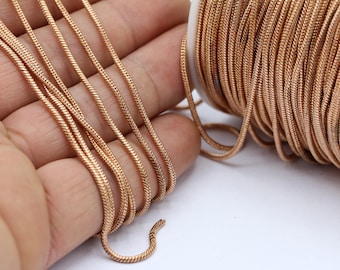 May include: A spool of gold-colored snake chain, a type of jewelry chain, is shown on a white surface. Several strands of the chain are laid out on a hand.