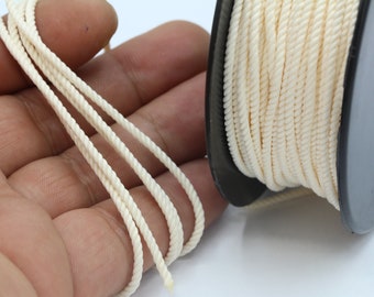 Può includere: Un rocchetto di cordoncino di cotone color crema, con diversi fili di cordoncino disposti su una mano.