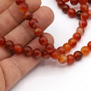 May include: Strands of round, polished beads in shades of red, orange, and brown. The beads have a banded pattern and are strung together, likely for jewelry making. The beads are held in a hand against a white background.
