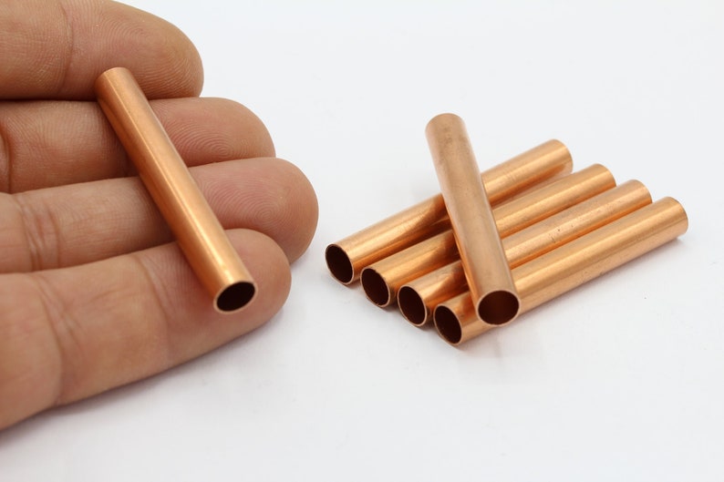 May include: Several copper-colored metal tubes, cylindrical in shape, are displayed. One tube is held in a hand, while the others are grouped together. The tubes have a smooth, reflective surface and open ends. These are likely craft or jewelry-making supplies.